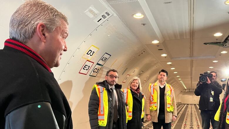 A man speaking to three people inside a cargo plane, a man with a camera stands to the right.