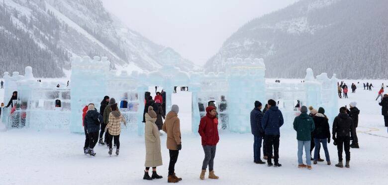 Dozens of bundled-up visitors tour around giant ice sculptures on the edge of Lake Louise on a snowy day, surrounded by mountains.