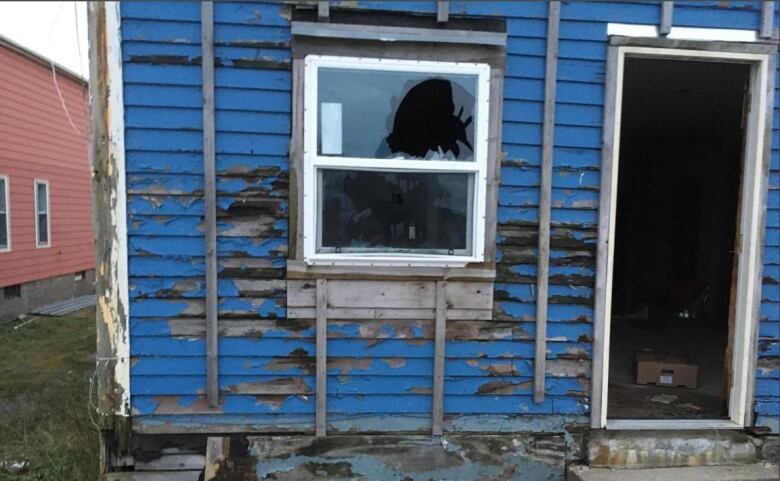 The rear view of a dilapidated house, showing signs of a break-in, with a busted window and wide-open rear door. 