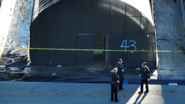 Three police officers in uniform stand next to a half-log shaped building with a partially open door in the middle. Police tape runs across the front of the building and 