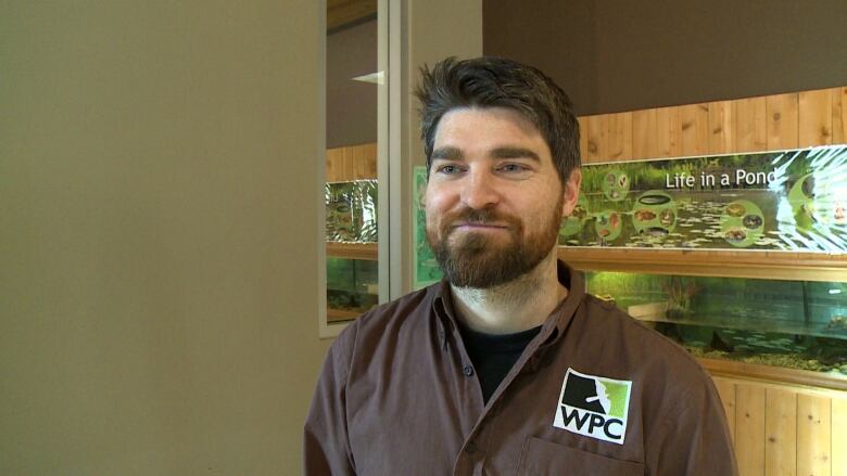 A man with brown hair and a beard wearing a brown shirt standing in front of an aquarium