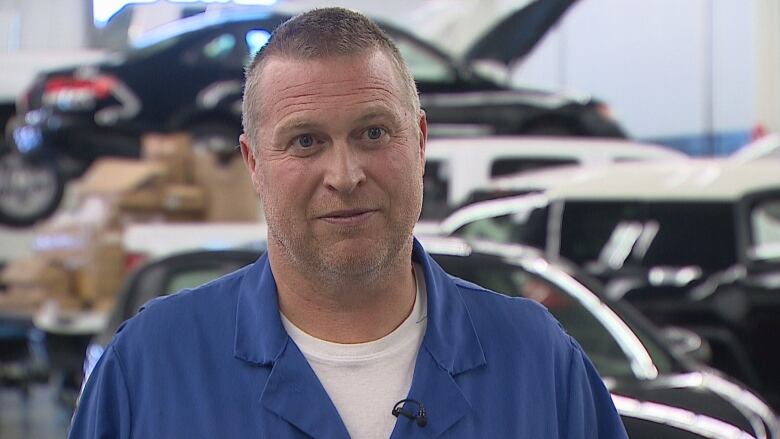 A white man with very short-cropped dark hair looks directly at the camera. He is smiling slightly and is wearing a white t-shirt underneath blue mechanic's overalls.