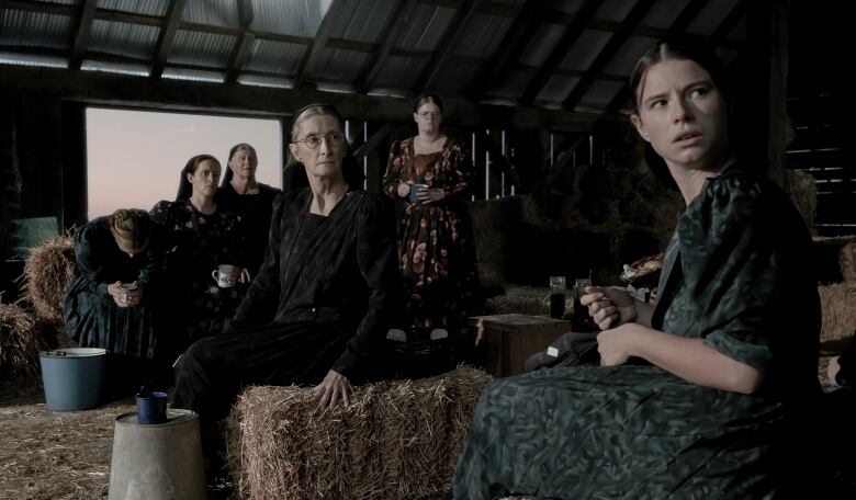 A group of women sit on bales of hay in a barn.