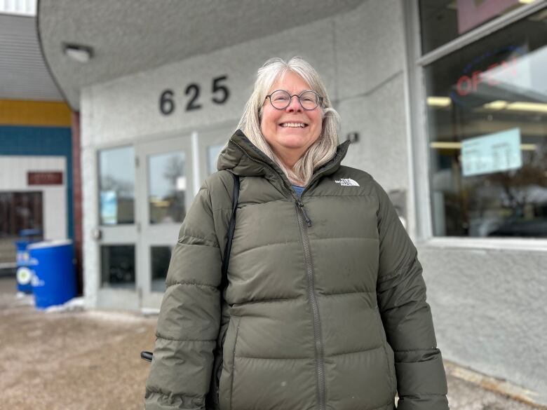 A woman in a parka is standing in front of a building and smiling at the camera.