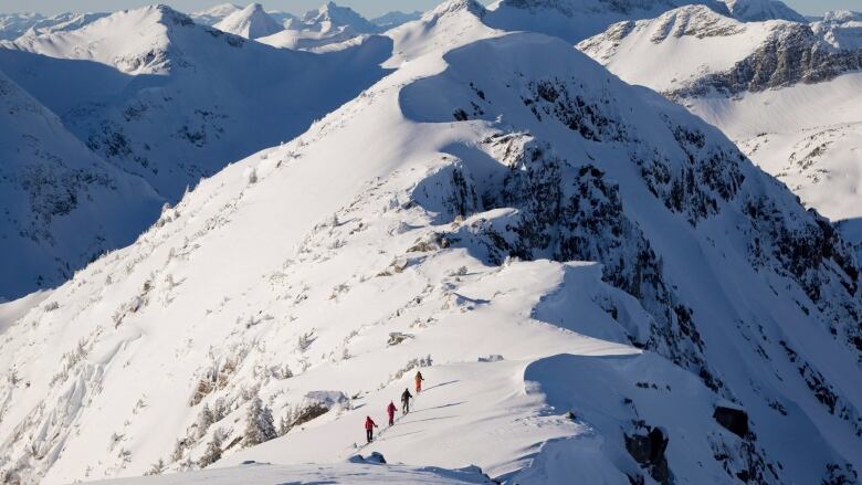 Backcountry skiers are dwarfed by the mountains as they make their way along a snowy mountain ridge.