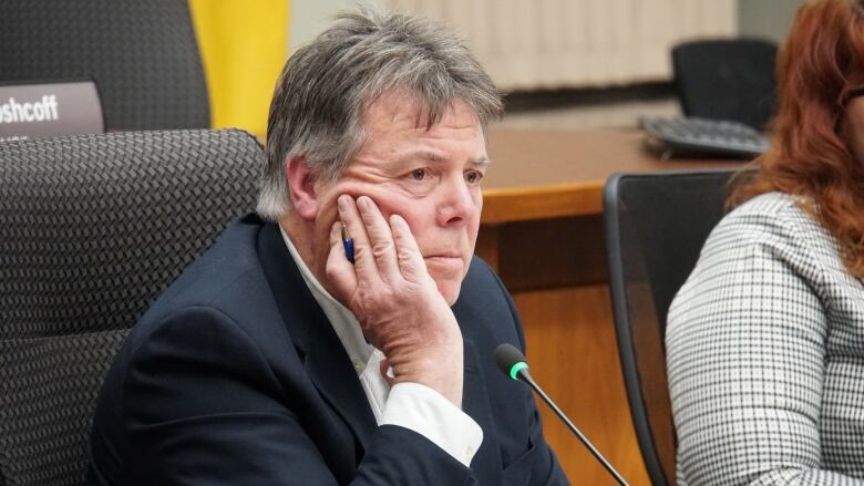 A pensive-looking man wearing a suit sits behind a microphone, his hand resting on his cheek.