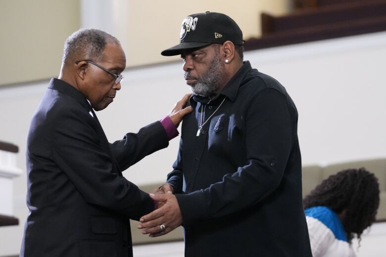 A man in religious attire shakes the hand of another man, dressed in black with a black ball cap on. 