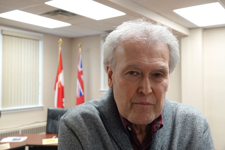 A man with white hair stares at the camera with a serious face. The flags of Ontario and Canada can be seen in the background behind him.