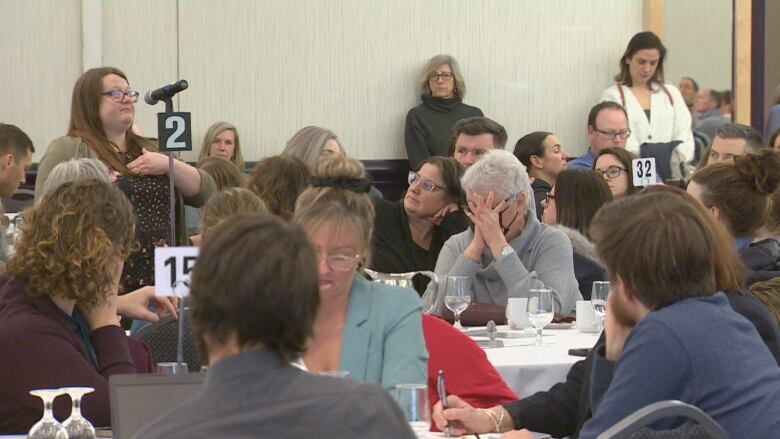 People sitting at round tables. A woman on the left is standing at a microphone