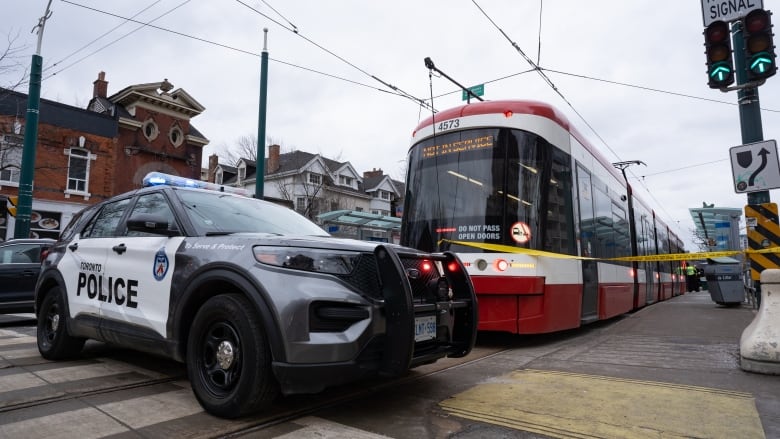 A grey police SUV is parked in front of a red and white streetcar with police tape on it.