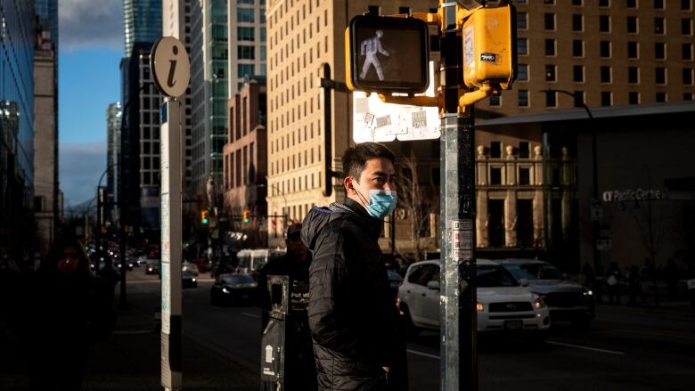 A man in a mask stands near a parking meter on a busy urban street flanked by highrises.