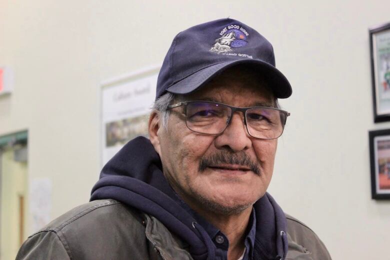 Head shot of a man wearing glasses and a blue baseball cap. You can also see he's wearing a jacket with a blue hoodie underneath it.