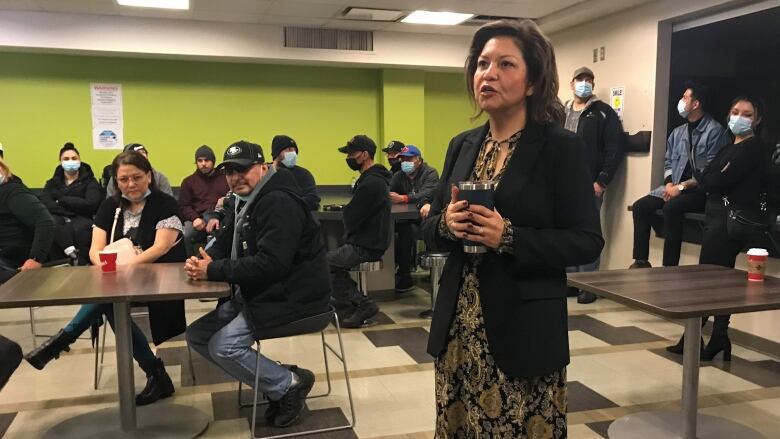 A woman is standing and speaking in a lime-green room with tiled-flooring. She is holding a thermos with both hands. Behind her is a cafeteria area, where many people are congregating.