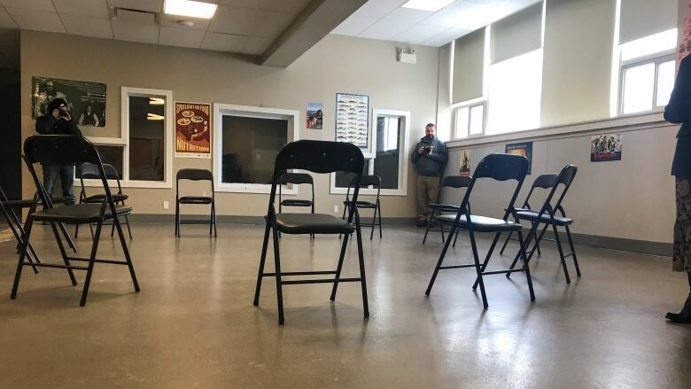 Foldable chairs are arranged in a circle in a beige-painted room, with several posters on the file. The room is lit by fluorescent lights, as well as natural light shining in through large windows. 