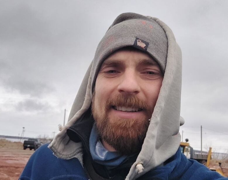 A man with a beard, wearing a hoodie and a toque, stands outside. 