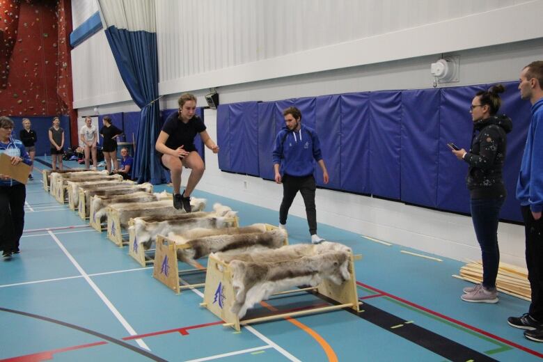 A teenage girl jumps over hurdles in a gym while coaches and other athletes watch.