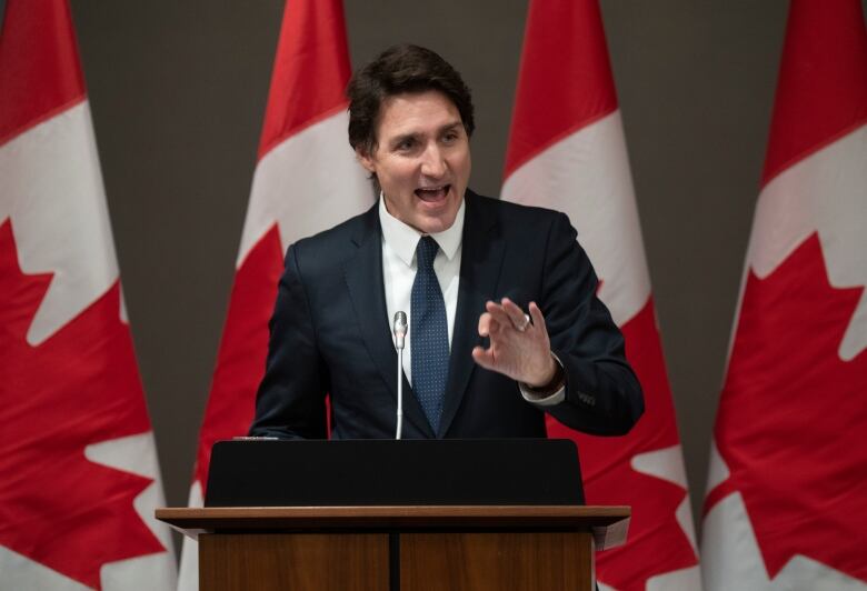 A man with brown hair and wearing a dark suit gestures with his hand as he speaks into a microphone at a podium.