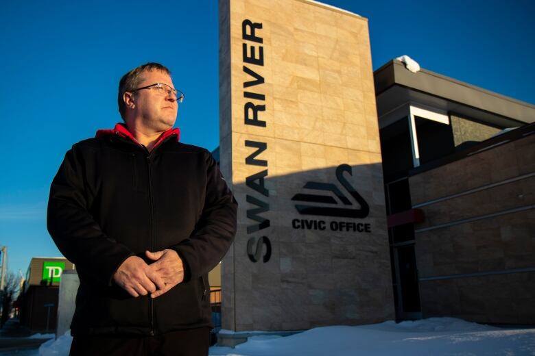 A man stands outside in front of a sign that says Swan River.