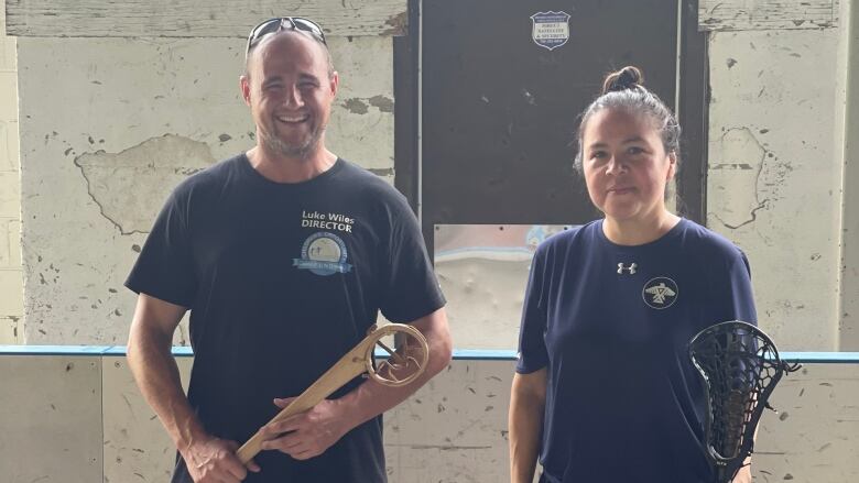 Two people stand holding various styles of lacrosse sticks.