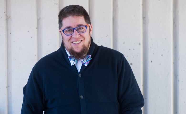 A bearded man, wearing glasses and a buttoned navy cardigan, stands in front of a wall. 