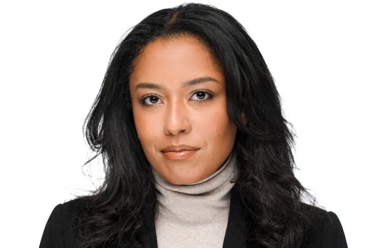 A woman with long, dark hair, wearing a grey turtleneck top and blazer, stands against a white background.