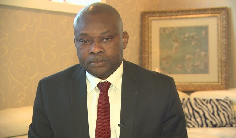 A man looks solemnly at the camera. He wears a black suit and red tie.