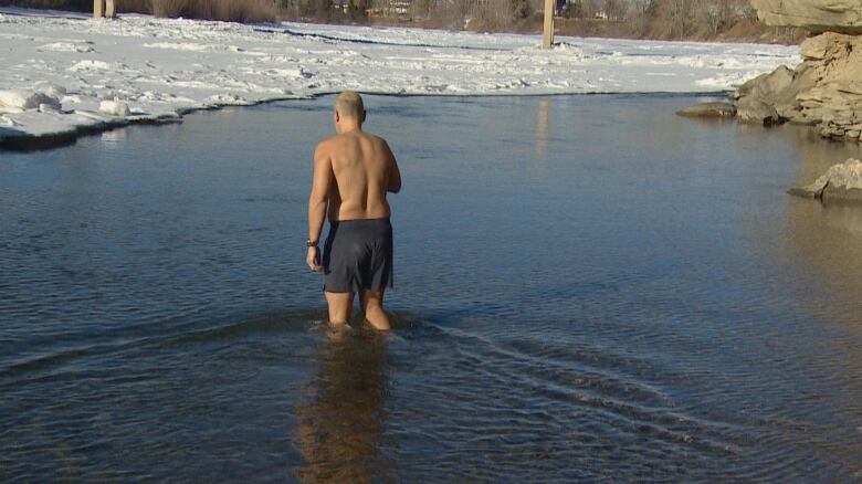 A man walking into the river with snow on the banks.