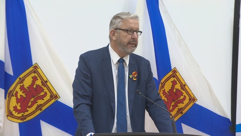 A man wearing a shirt in blazer stands in front of two Nova Scotia flags.