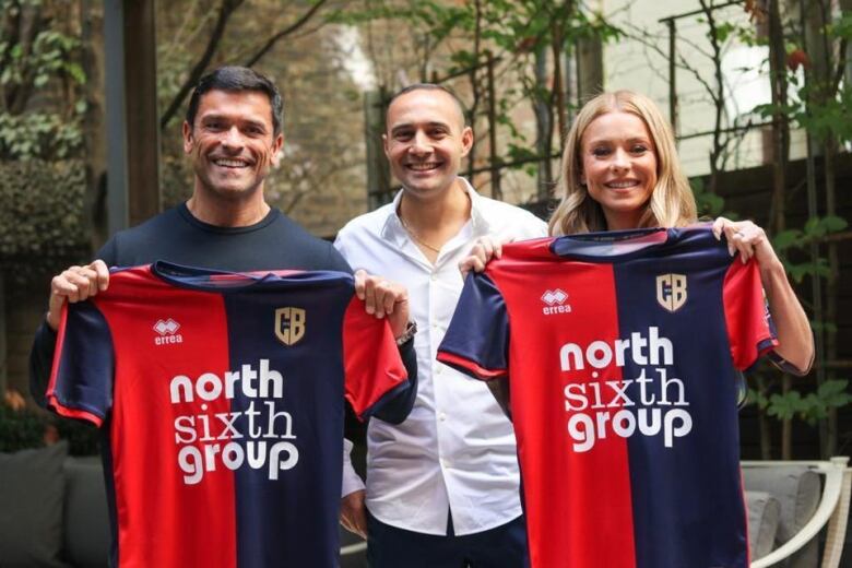 Smiling man and woman holding up soccer jerseys. They flank a smiling man dressed in white.