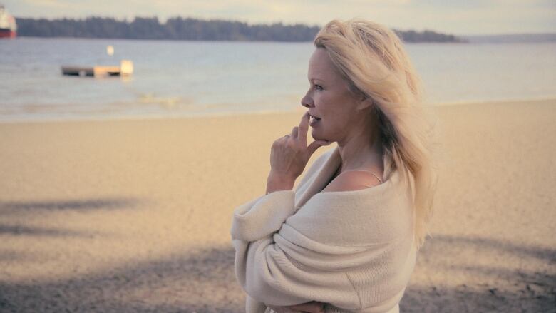 Blonde woman gazes into the distance as she stands on a bright sandy beach in front of open waters