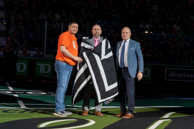 Two men stand around a third in the centre with a black and white star blanket wrapped around him