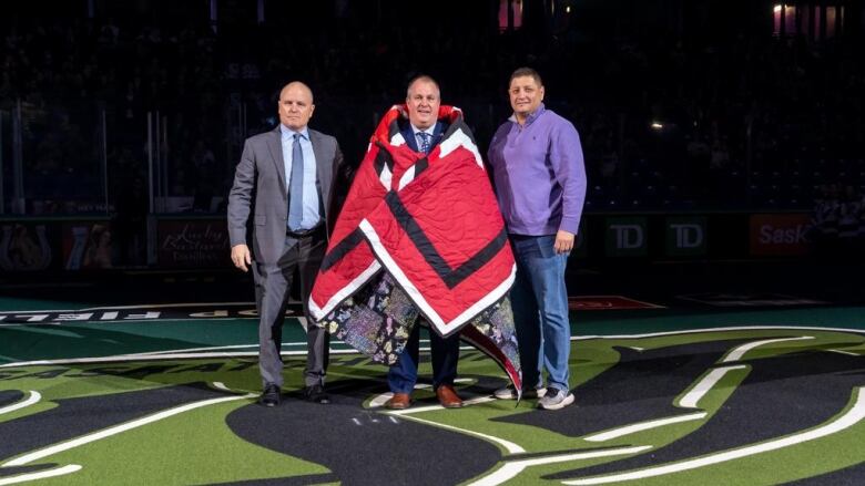 Three men stand in the centre of a stadium, one with a red, black and white star blanket wrapped around him.