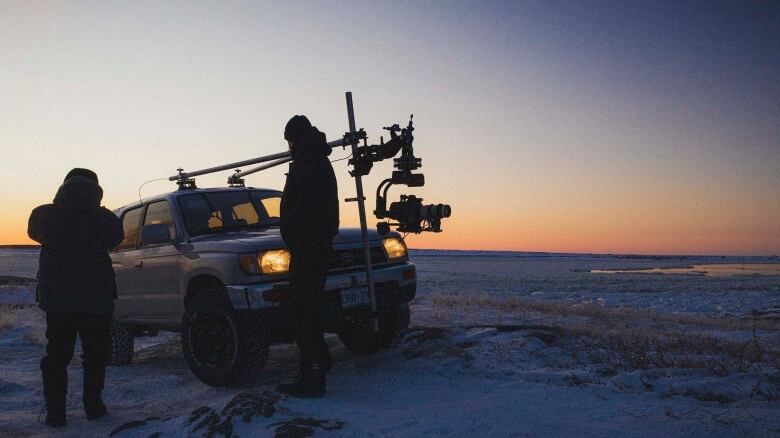 Two people stand in front of a large van with a camera rig attached to the front in a snow-covered landscape at dusk.