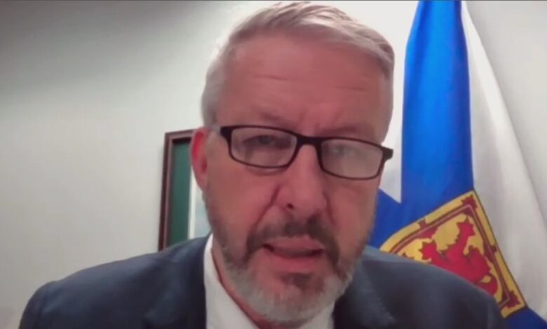 A man wearing a gray blazer and white shirt sits in front of the Nova Scotia flag.