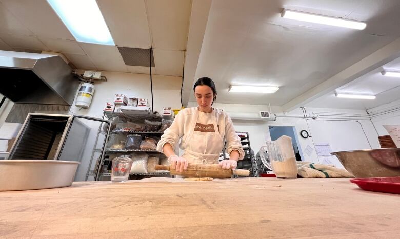 A woman with her dark hair pulled away from her face rolls dough on a crowded wooden surface.