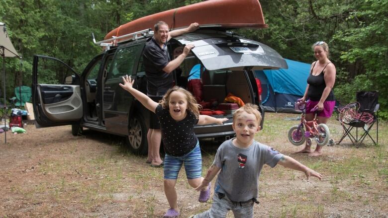 Two children run toward the photographer with big smiles on their faces as a man and woman, presumably their parents, unpack a van with camping gear.