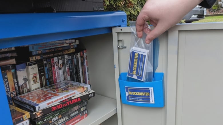 An outdoor blue cabinet is open, displaying DVDs, VHS tapes, and a stack of Blockbuster membership cards.