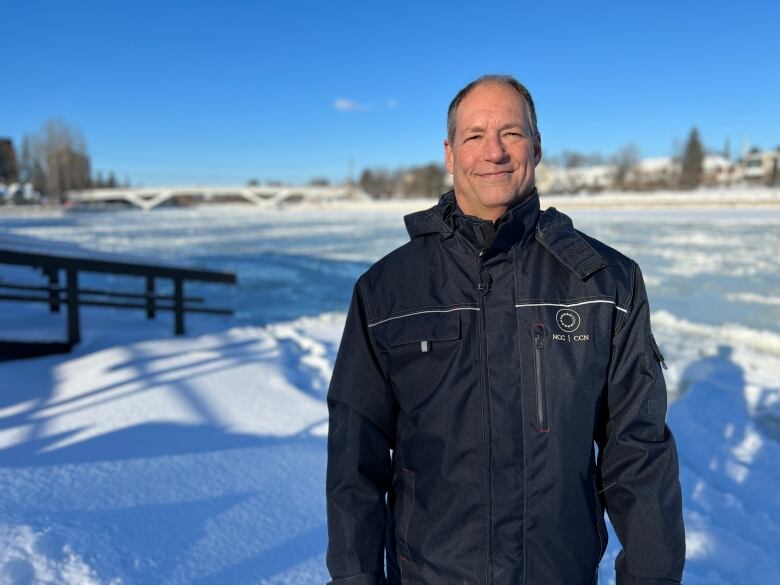 A man in a jacket stands in front of a snowy, icy body of water on a clear winter day.