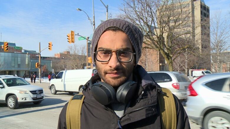 A man wearing a grey hat, glasses, a brown backpack and a black jacket with headphones around his neck