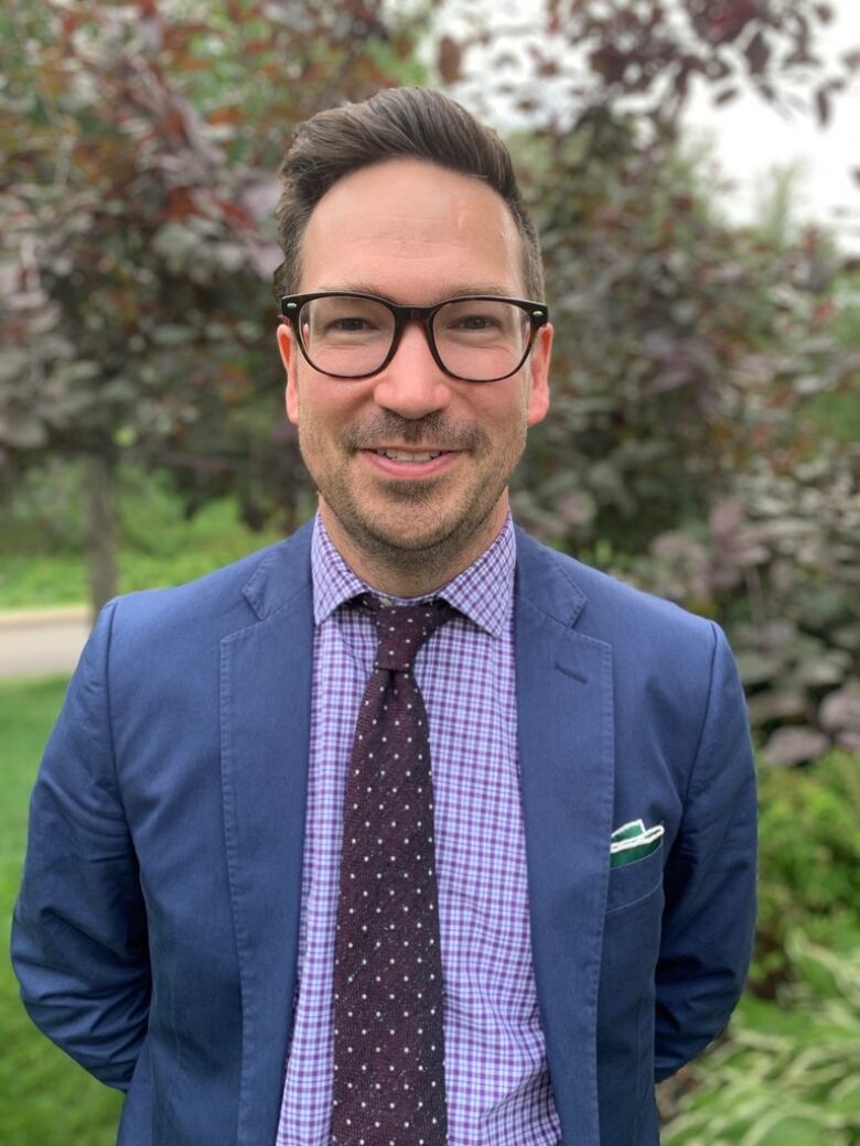 A man in a blue suit with a purple checker shirt and dark purple tie smiles at the camera while standing outside.