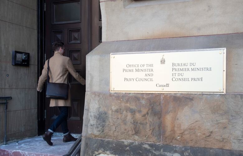 A person in a tan coat walks in the wooden door of a large brick building that has a plaque on the exterior wall.