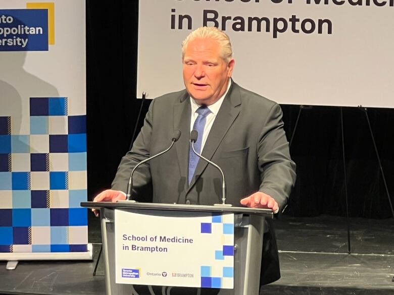 A white man stands at a podium with the words 'School of medicine in Brampton' written on it. He is wearing a suit with a blue tie.