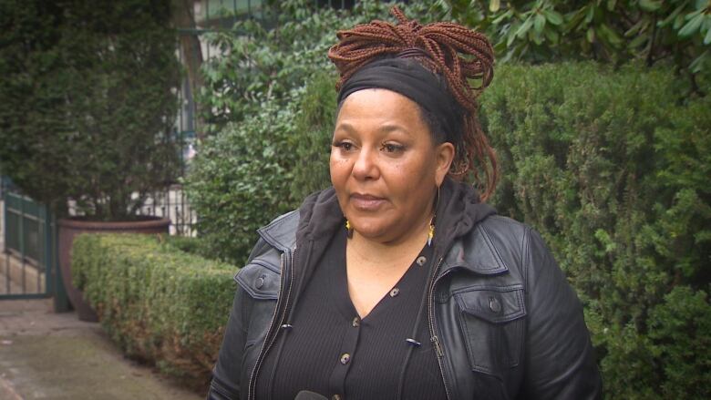 A woman with her hair in a black bandana, wearing a black leather jacket over a black open-neck sweater, stands in front of a green hedge.