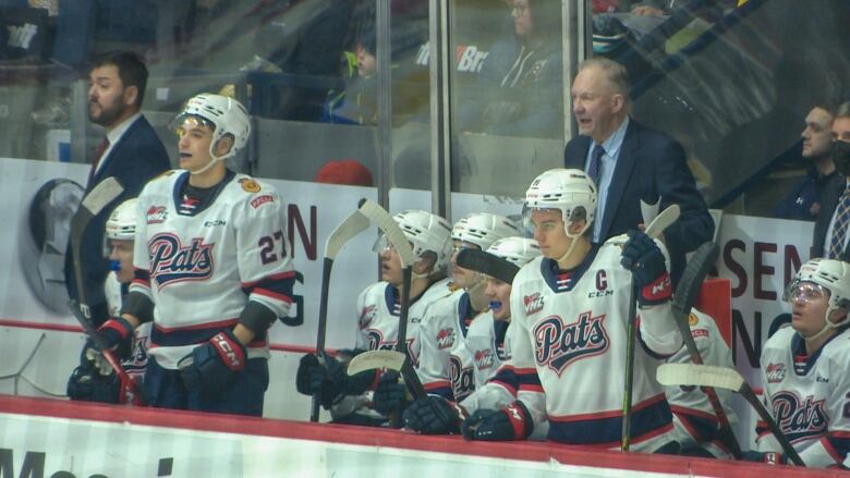 Hockey players are on the bench. Two of the players are standing up. The head coach, dressed in a suit and tie, is shouting.