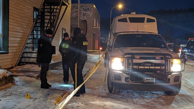 Three police investigators behind police tape and a pickup truck near a building.