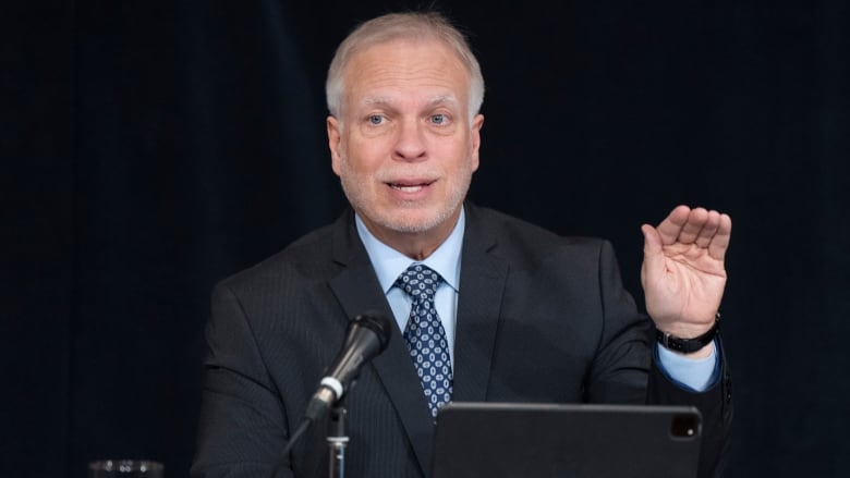 Man in dark suit talks into a mic with one hand raised.