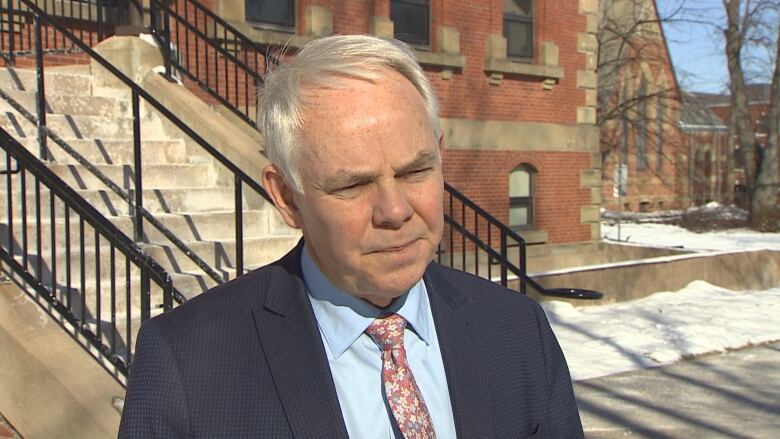 Man with white hair wearing blue shirt, flowery tie and navy jacket stands in front of brick building.