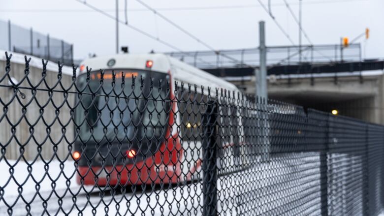 A red-and-white train travels along snowy tracks behind a chain-link fence.