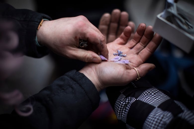 A closeup photo of a hand shows a pile of blue steel-like pills and purple pills in small zip-lock bags.