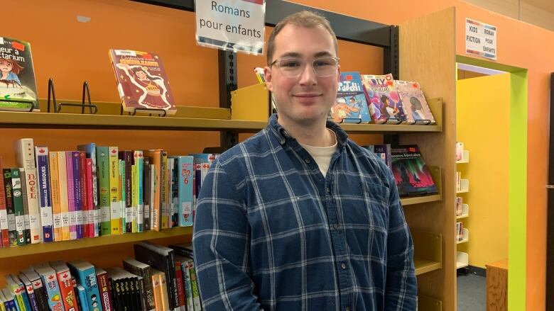 A young man with glasses in a plaid shirt standing in a library.